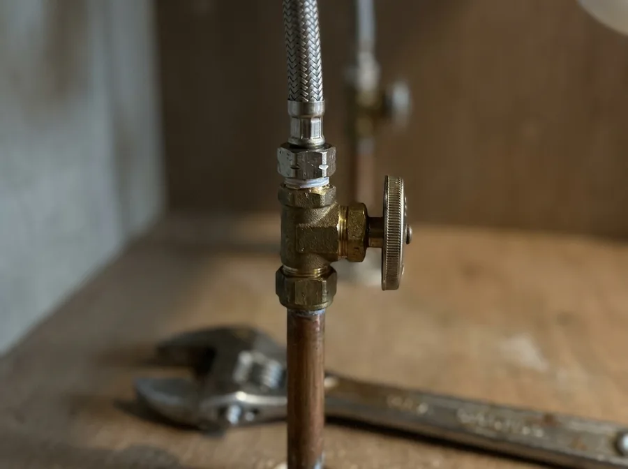 very close crop under a kitchen sink on a repaired angle stop connected to the riser below and a braided supply line above, a wrench resting on the dry cabinet floor, shallow depth of field